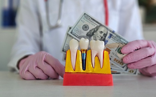 Gloved hands holding $100 behind large model dental implant